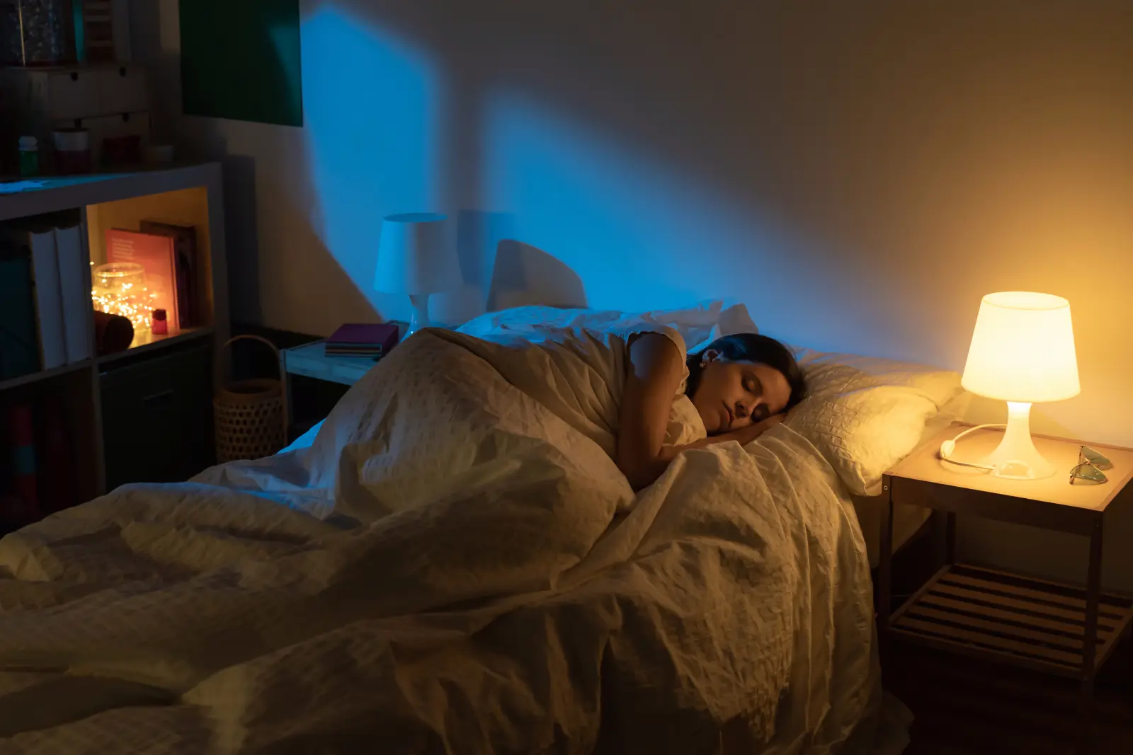 Woman sleeping in bed with a bedside lamp and bookshelf light on