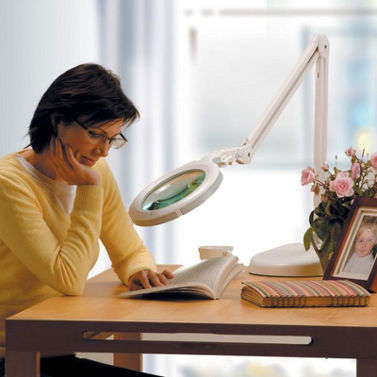 Woman sitting at the table reading a book through large adjustable magnifying lamp.